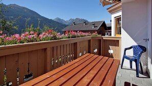 Una terraza de madera con un banco y una silla, con vistas a un paisaje montañoso que incluye una construcción de madera y flores rosadas en primer plano.