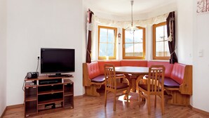 Una sala de estar con televisor, mueble de entretenimiento de madera, un banco tapizado rojo con una mesa y sillas.
