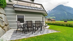 Une maison moderne dotée d’un patio comprenant une table et des chaises, le tout entouré d’une pelouse bien entretenue et avec une chaîne de montagnes en toile de fond.