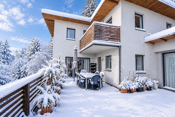 A modern two-story house with a balcony, a dining area with chairs and a table, and snow-covered trees and ground.