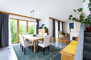 A modern living space with a dining area, a sofa, a staircase, and a view of greenery outside.