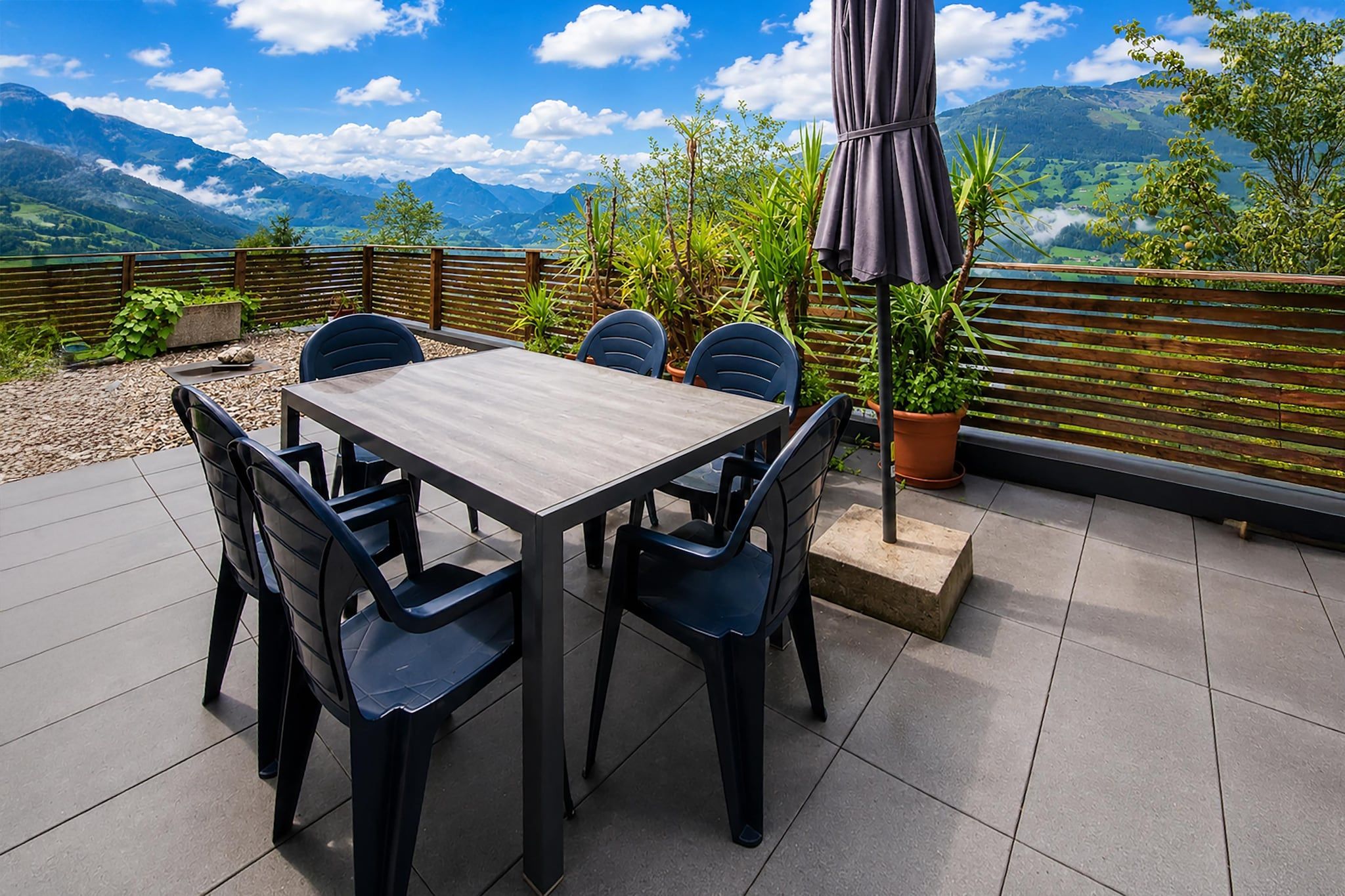 Een terras buiten met een tafel en stoelen, omringd door planten en uitzicht op bergen.