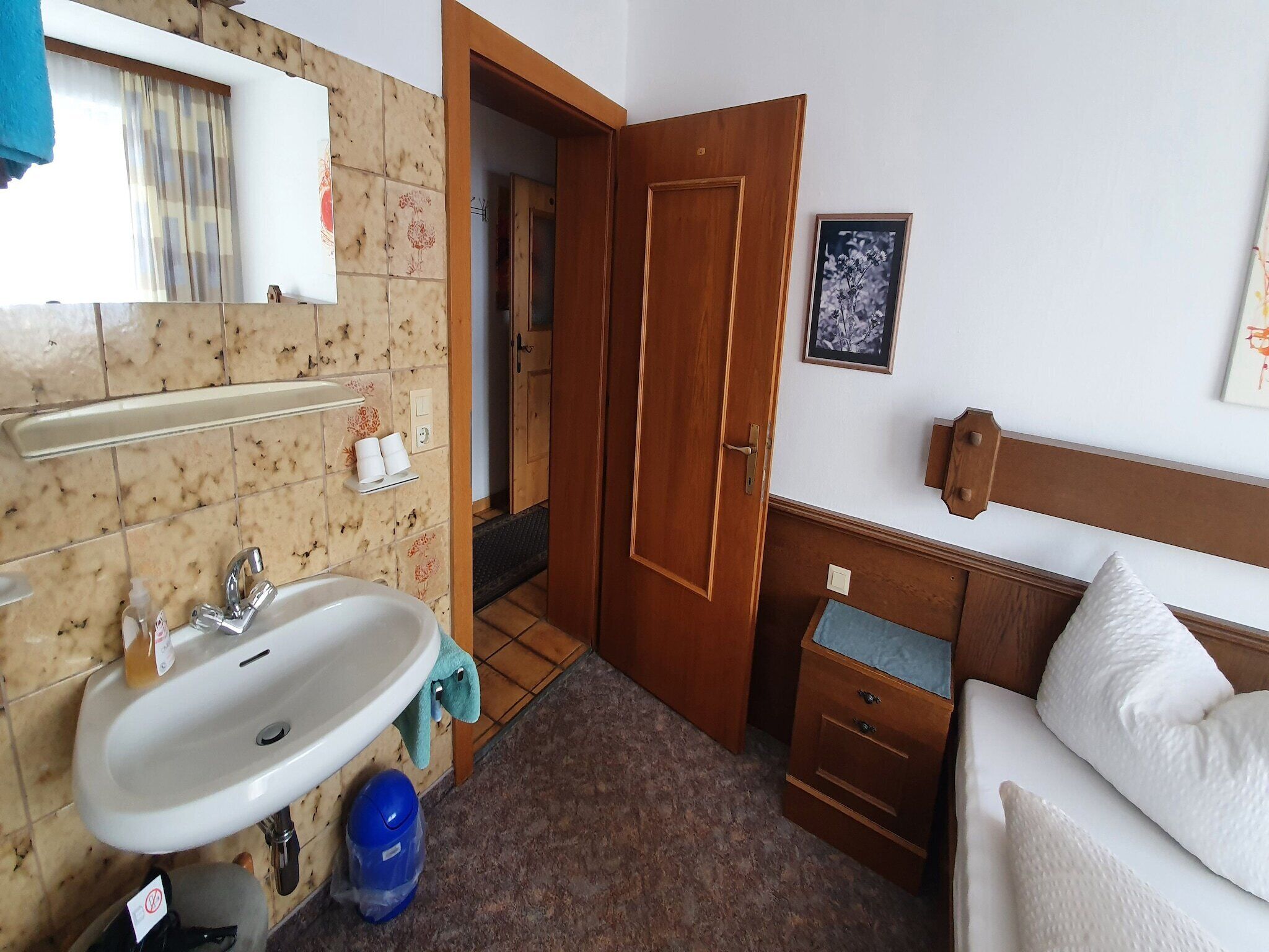 A hotel room with a sink, wooden door, bedside table, and a bed with pillows.