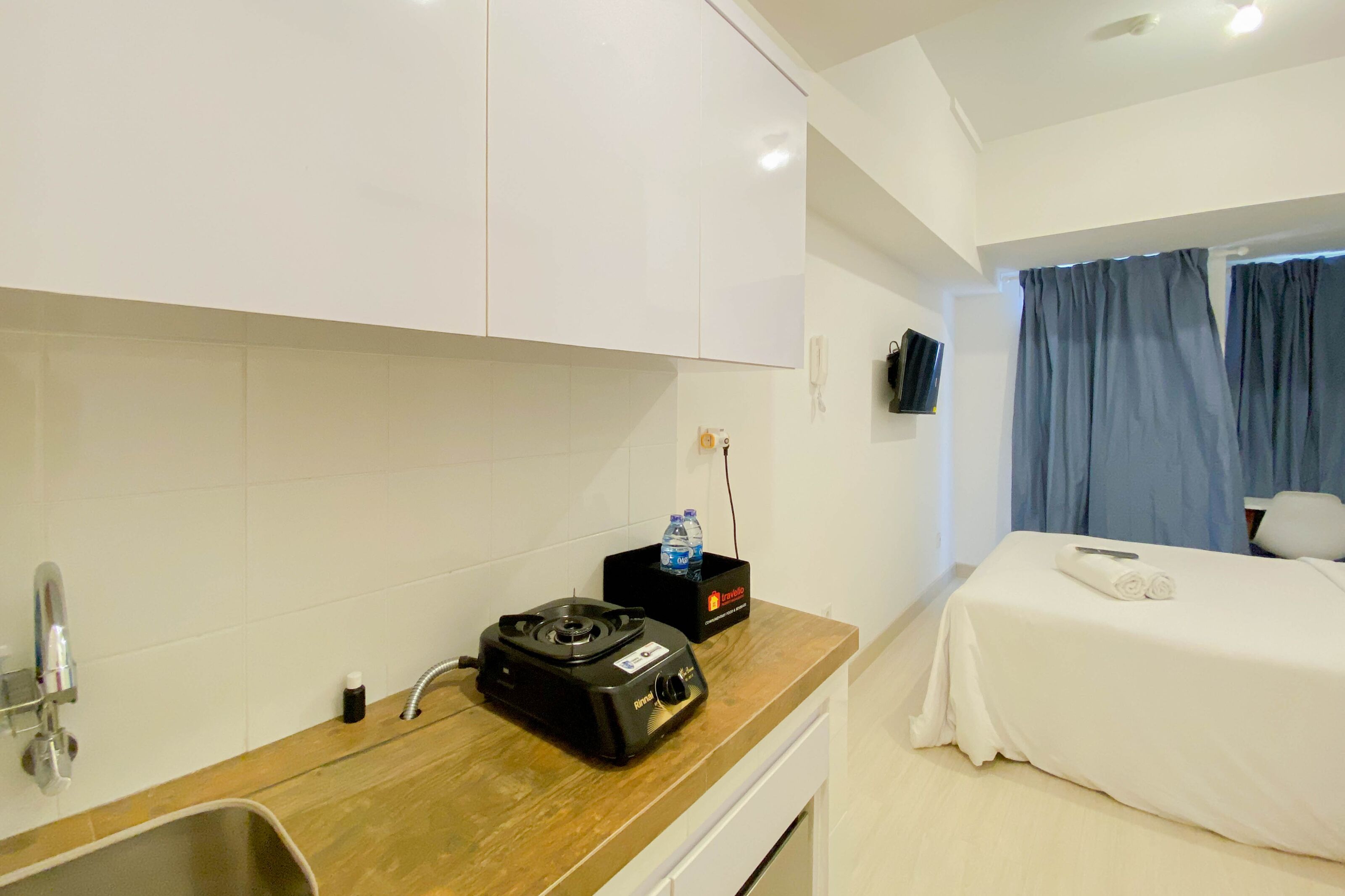 A hotel room with a kitchenette, a bed with white bedding, a TV mounted on the wall, and a window with blue curtains.