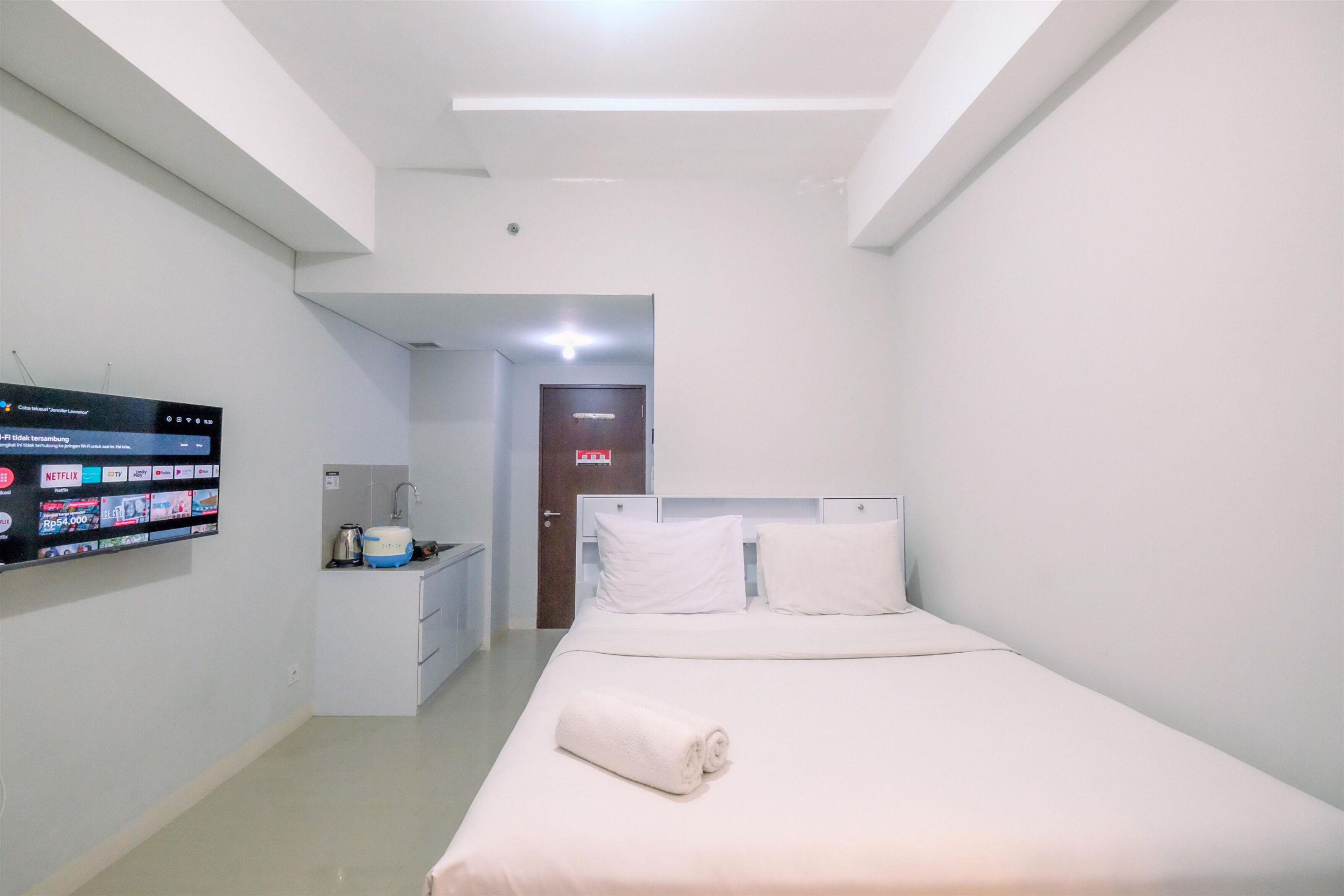 A hotel room with a bed, a TV mounted on the wall, a small kitchenette, and a door leading to another room.