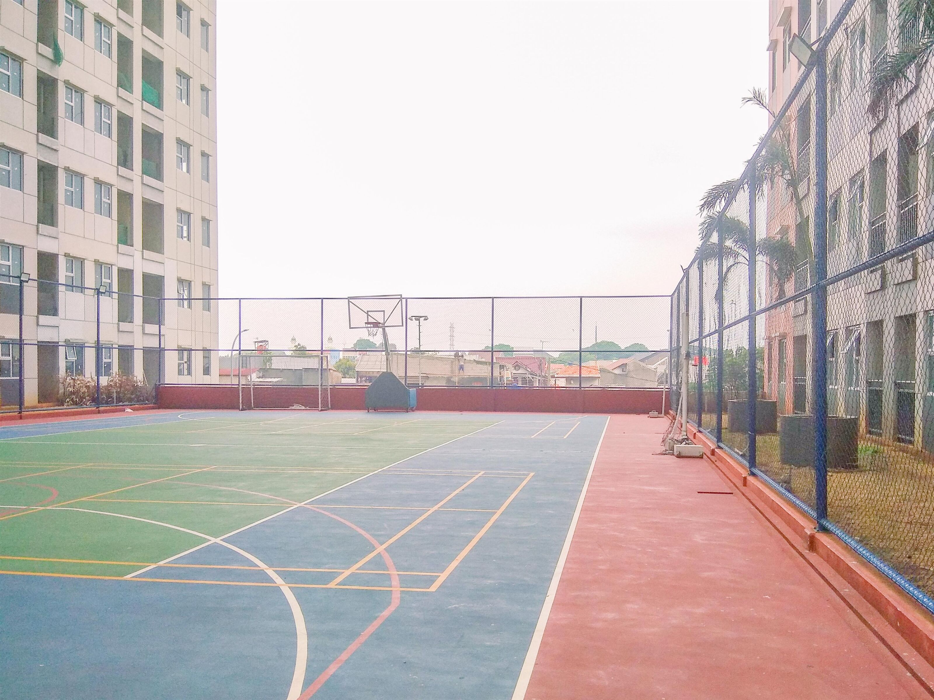 An outdoor basketball court with a red and blue playing surface, surrounded by a chain-link fence.