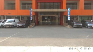 Un edificio moderno con fachada roja y naranja, un acceso cubierto y un estacionamiento con varios automóviles.
