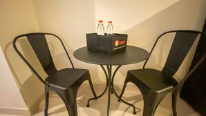 A small round table with two black chairs, a box labeled 'Travelio' with two bottles, and a plain wall in the background. - Minimalist Studio Room Marquis De Lafayette Apartment (Semarang)