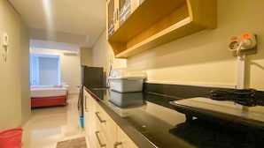 A modern kitchen with a black countertop, white cabinets, and a wooden shelf above. There is a red chair in the background and a small red basket on the floor. - Minimalist Studio Room Marquis De Lafayette Apartment (Semarang)