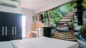 A hotel room with a bed, a TV mounted on the wall, a wardrobe, a small kitchenette, and a mural of a forest scene. - Warm And Cozy Stay Studio Green Bay Pluit Apartment (Jakarta)