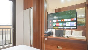 Une chambre d’hôtel dotée d’un grand téléviseur à écran plat fixé au mur, qui diffuse différents services de streaming. On y trouve un bureau en bois avec un ordinateur et une bouteille d’eau. La chambre est équipée d’un lit avec une literie blanche et d’une fenêtre donnant sur l’extérieur.