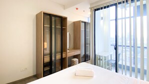 A hotel room with a large bed, a wardrobe, a small table with a lamp, and a view of the city through large windows. - Elegant And Comfort 1Br Sudirman Suites Apartment (Jakarta)