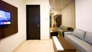 A modern living room with a flat-screen TV mounted on the wall, a dark door, a staircase, and a grey sofa. - Elegant And Comfort 1Br Sudirman Suites Apartment (Jakarta)