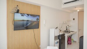 Kompakti oleskelutila, jossa on seinään kiinnitetty televisio puupaneeliseinässä, pieni keittiö jääkaapilla sekä juomavesiautomaatti.