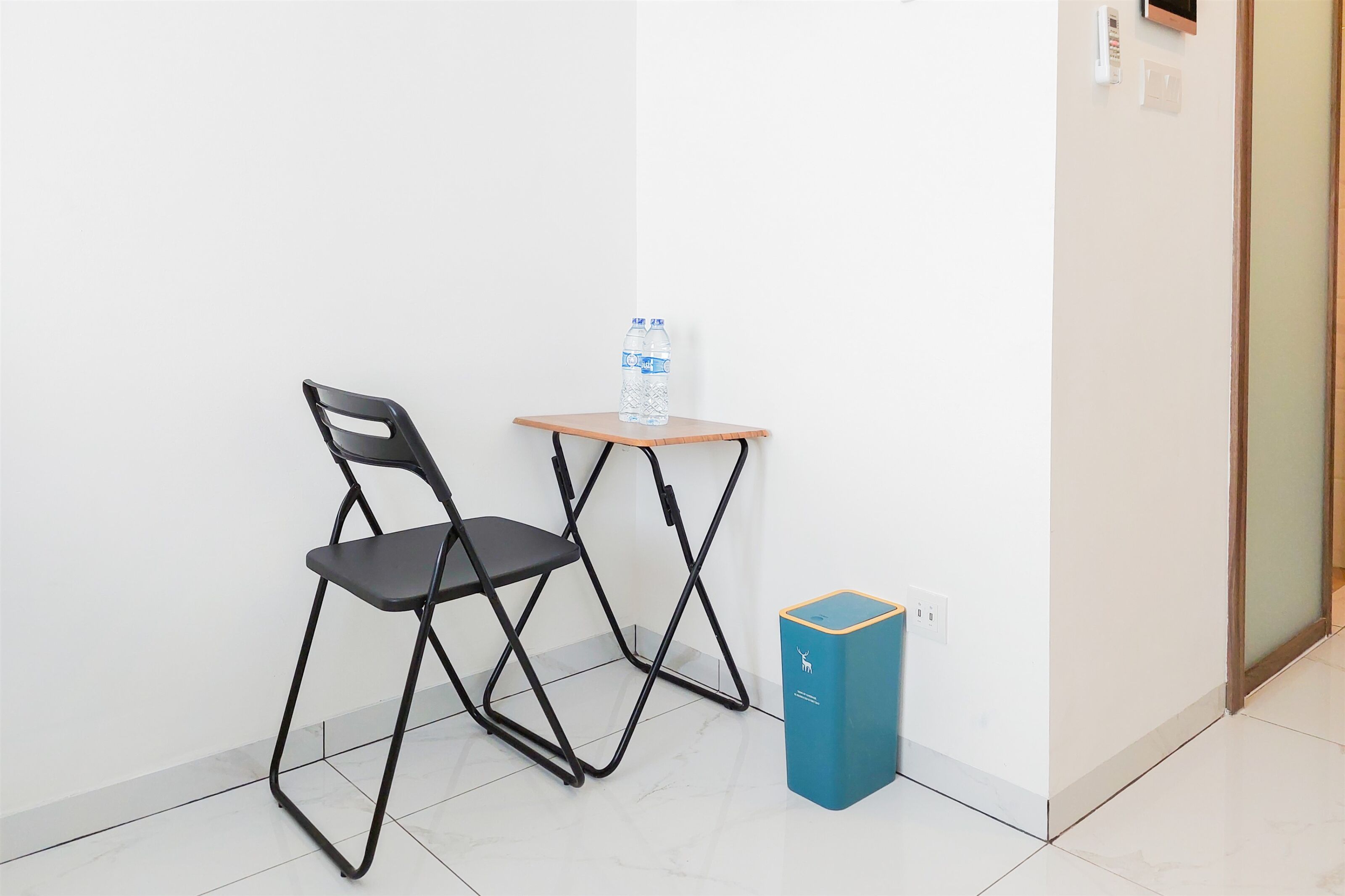 Un coin avec une chaise pliante noire, une petite table en bois sur laquelle se trouve une bouteille d’eau, et une poubelle bleue.
