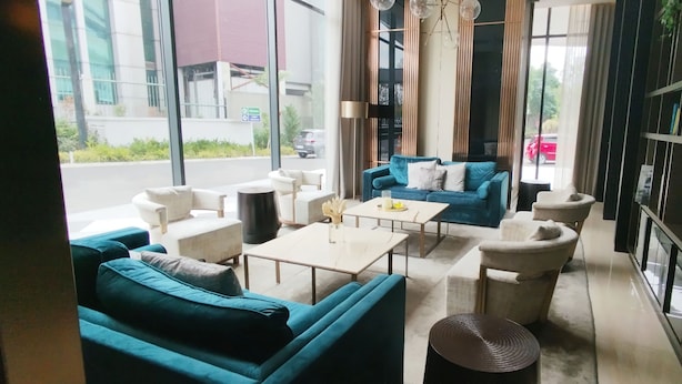 A modern hotel room with a blue sofa, a white armchair, a small table, and a large window offering a view of the street.