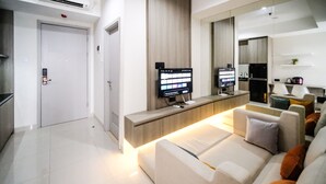 A modern living room with a sofa, a coffee table, and a TV mounted on the wall. There is a door and a kitchen area visible in the background. - Good Choice And Homey 1Br Grand Sungkono Lagoon Apartment (Surabaya)