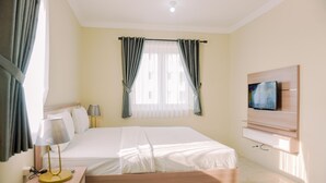 A hotel room with a bed, bedside table with lamp, TV mounted on wall, and curtains on the window. - Comfortable And Modern 2Br At Grand Palace Kemayoran Apartment (Jakarta)