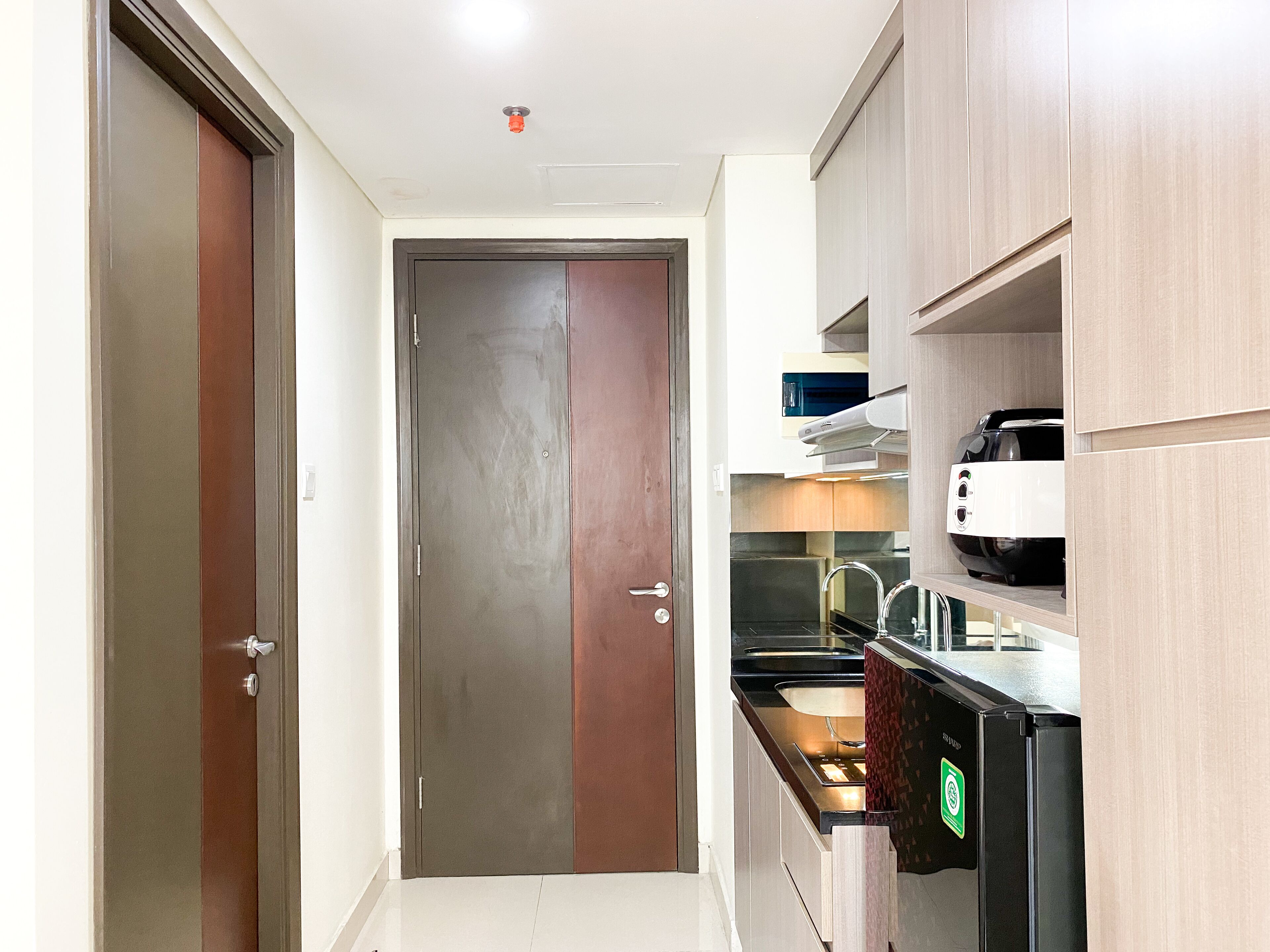 A narrow hallway leading to a door with a kitchen area on the right, including a stove, sink, and a black refrigerator.