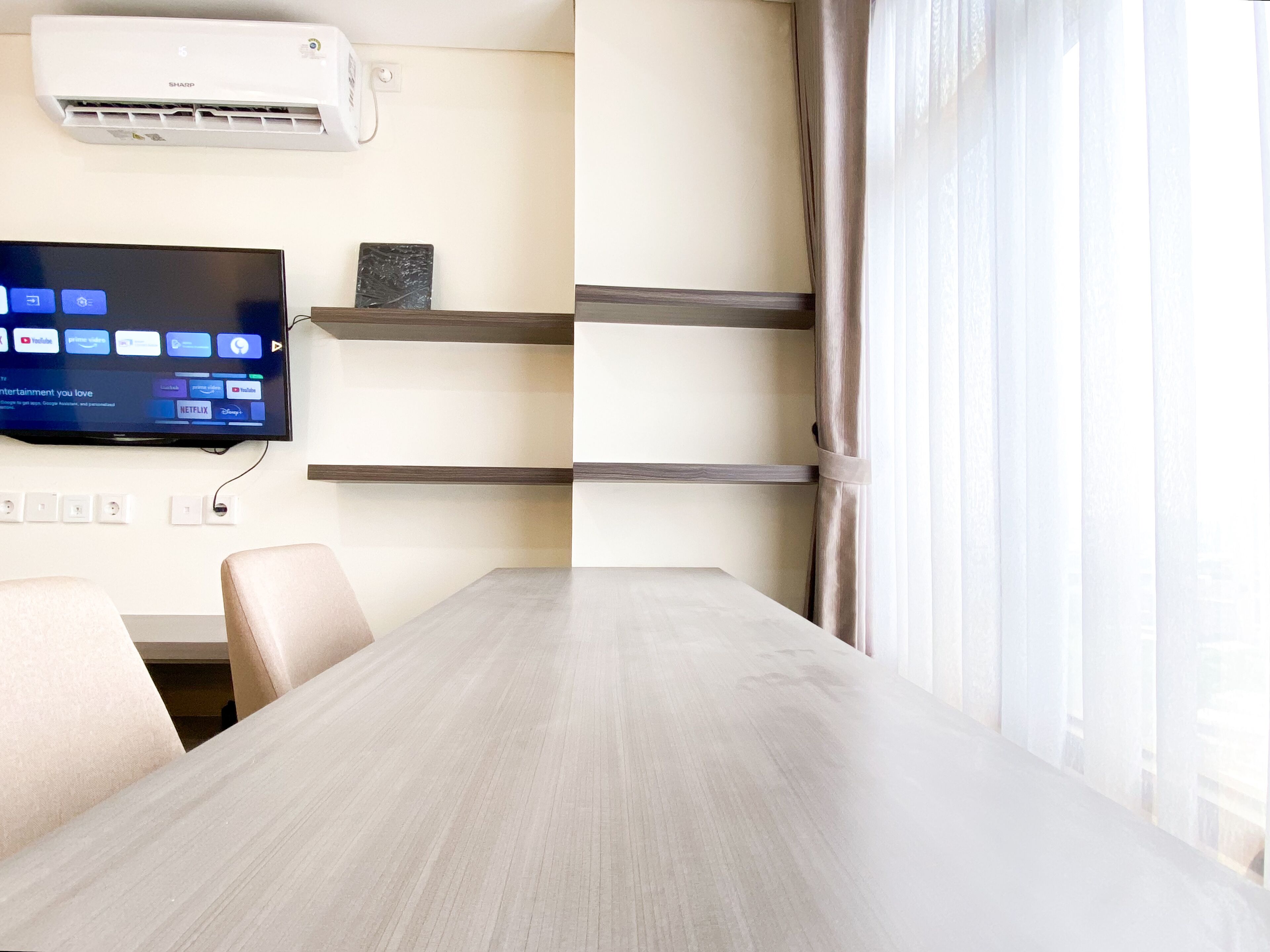 A modern hotel room with a wooden dining table, beige chairs, a mounted flat-screen TV displaying streaming services, a wall-mounted air conditioner, and a window with sheer curtains.