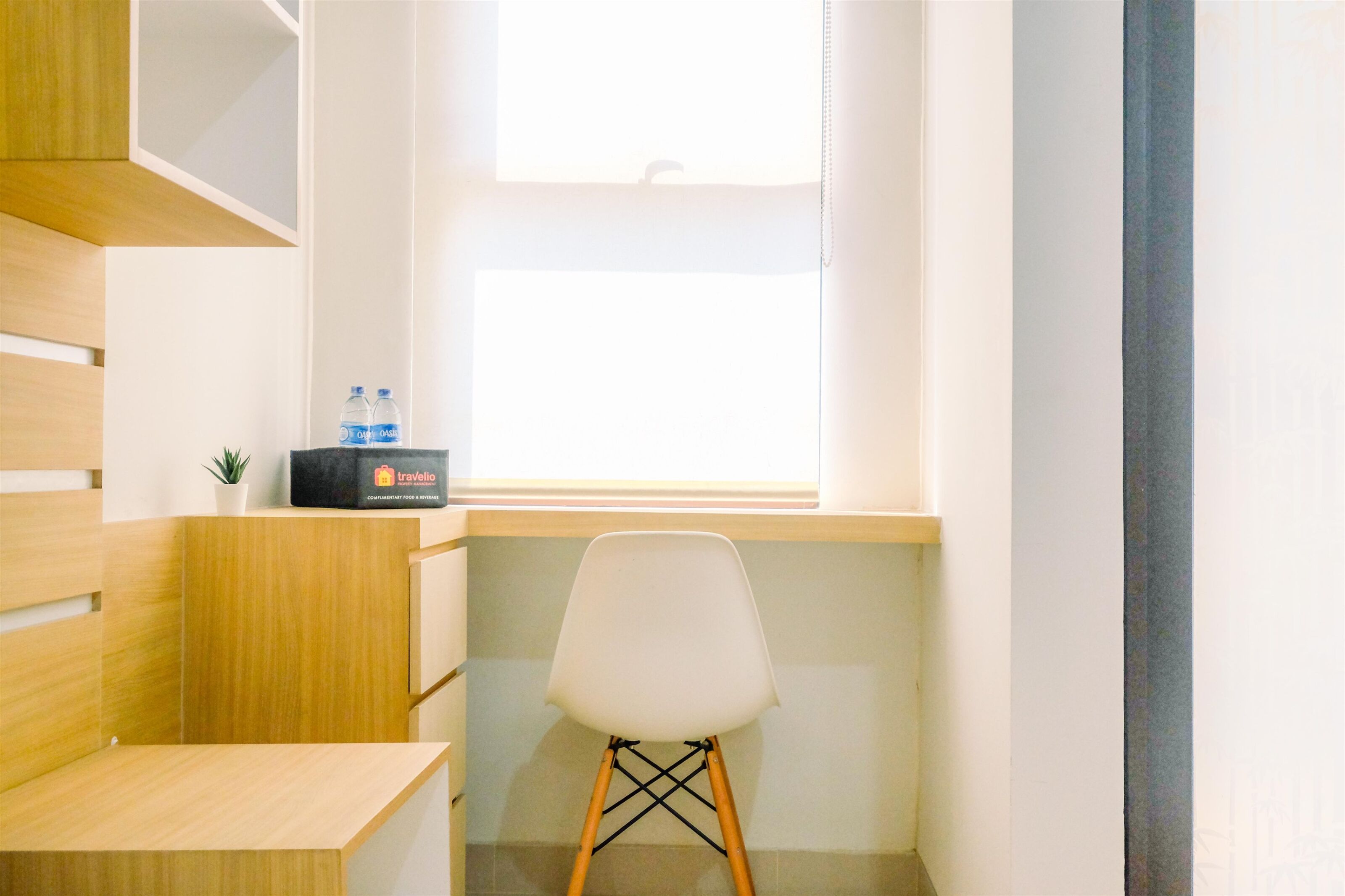 A small, modern office space with a wooden desk, a white chair, a small potted plant, and a window with natural light.