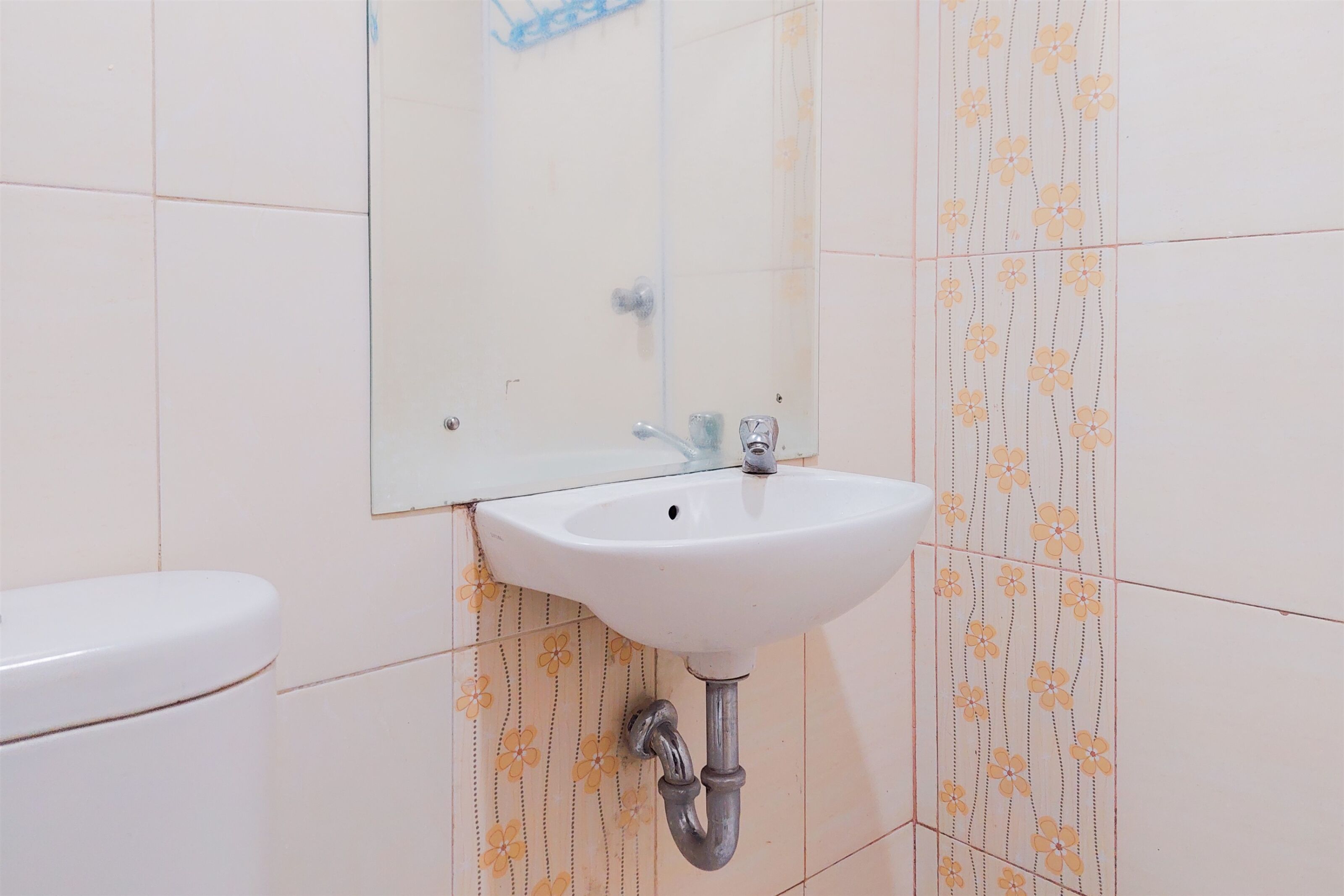 Un baño con lavamanos blanco, un espejo y un inodoro. Las paredes presentan un diseño de azulejos con motivos florales.