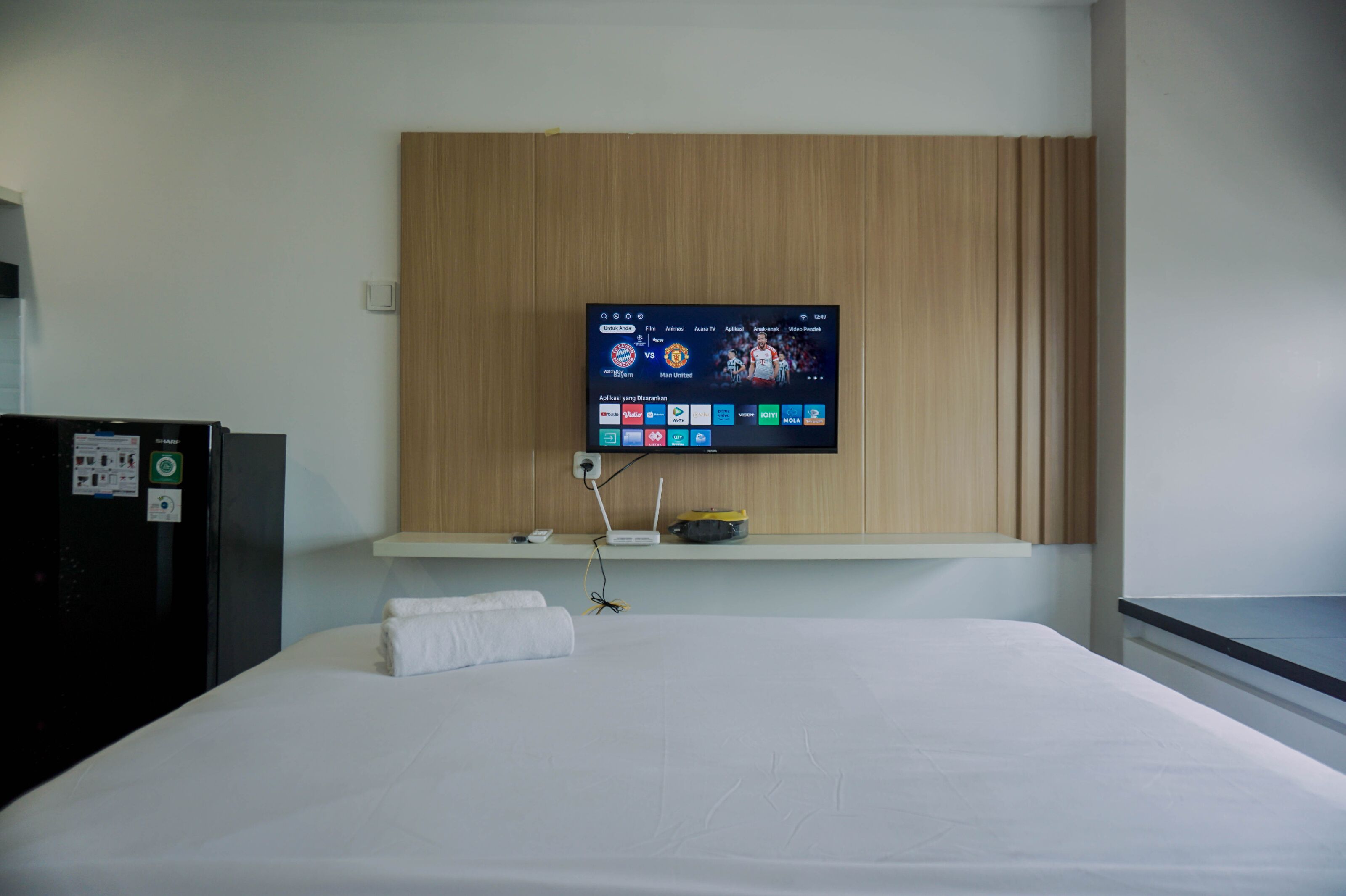 A hotel room with a bed, a TV mounted on a wooden panel, a small shelf with a remote control, and a refrigerator.