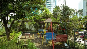 Un terrain de jeux avec des balançoires et un petit abri, entouré d’arbres et de plantes.