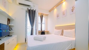 A hotel room with a bed, a desk with a computer, and a window with curtains. - Good And Homey Studio At Transpark Cibubur Apartment (Depok)