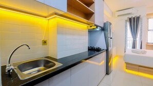 A modern kitchen with a stainless steel sink, a gas stove, and a built-in oven. There is a wooden shelf above the stove and a window with curtains. - Good And Homey Studio At Transpark Cibubur Apartment (Depok)