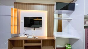A modern living room with a flat-screen TV mounted on a wooden wall, a built-in shelf, and a small green trash bin. - Scenic Studio Room Apartment Vida View Makassar (Makassar)