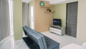 A modern living room with a grey sofa, a flat-screen TV on a wooden stand, and a door with a grey shutter. - Best Deal And Modern 2Br Amazana Serpong Apartment (South Tangerang)