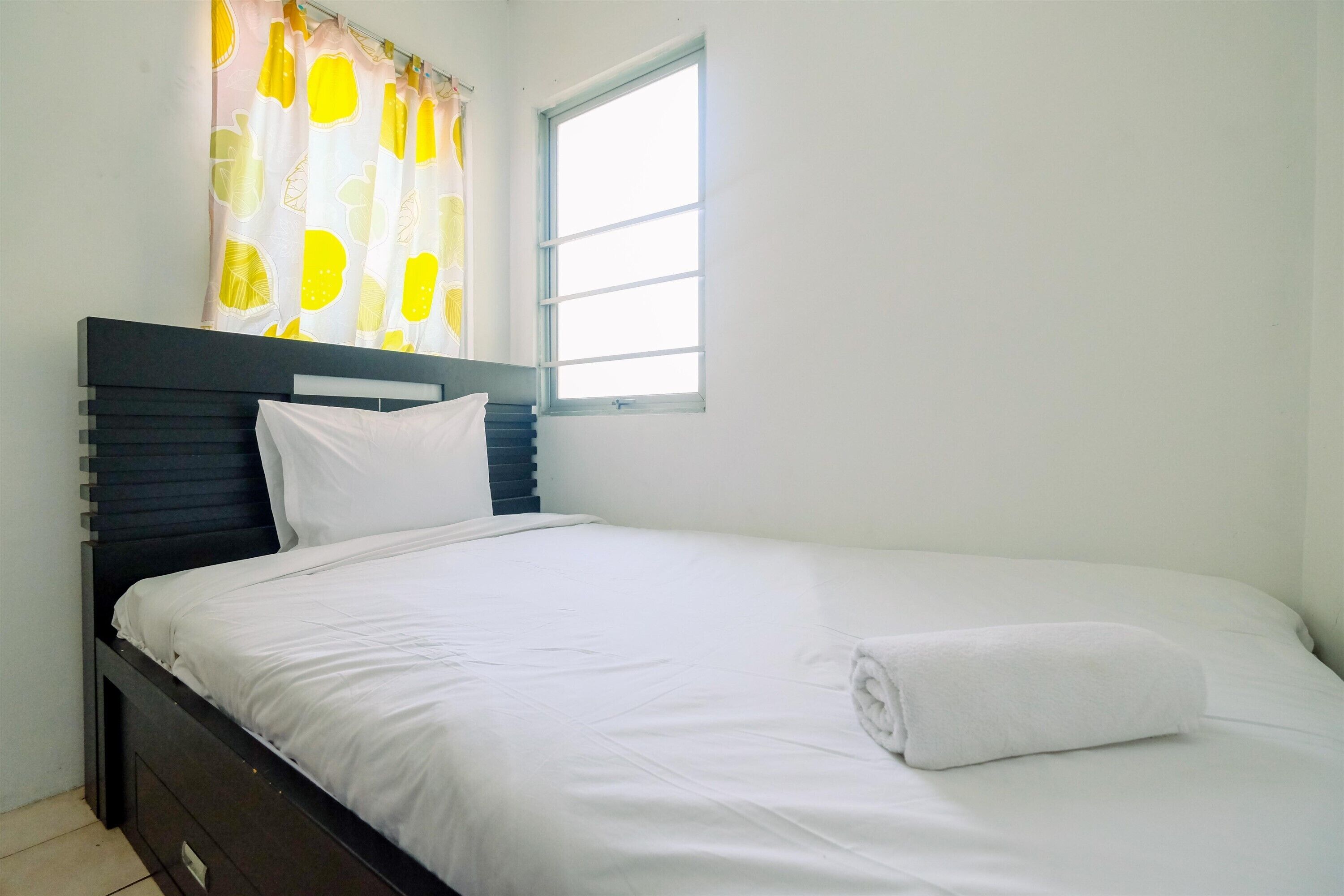 A neatly made bed with white linens and a rolled towel, a dark headboard, and a window with a white frame.