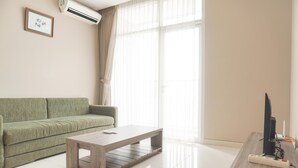 A living room with a green sofa, a wooden coffee table, a TV on a wooden stand, and a large window with sheer curtains.
