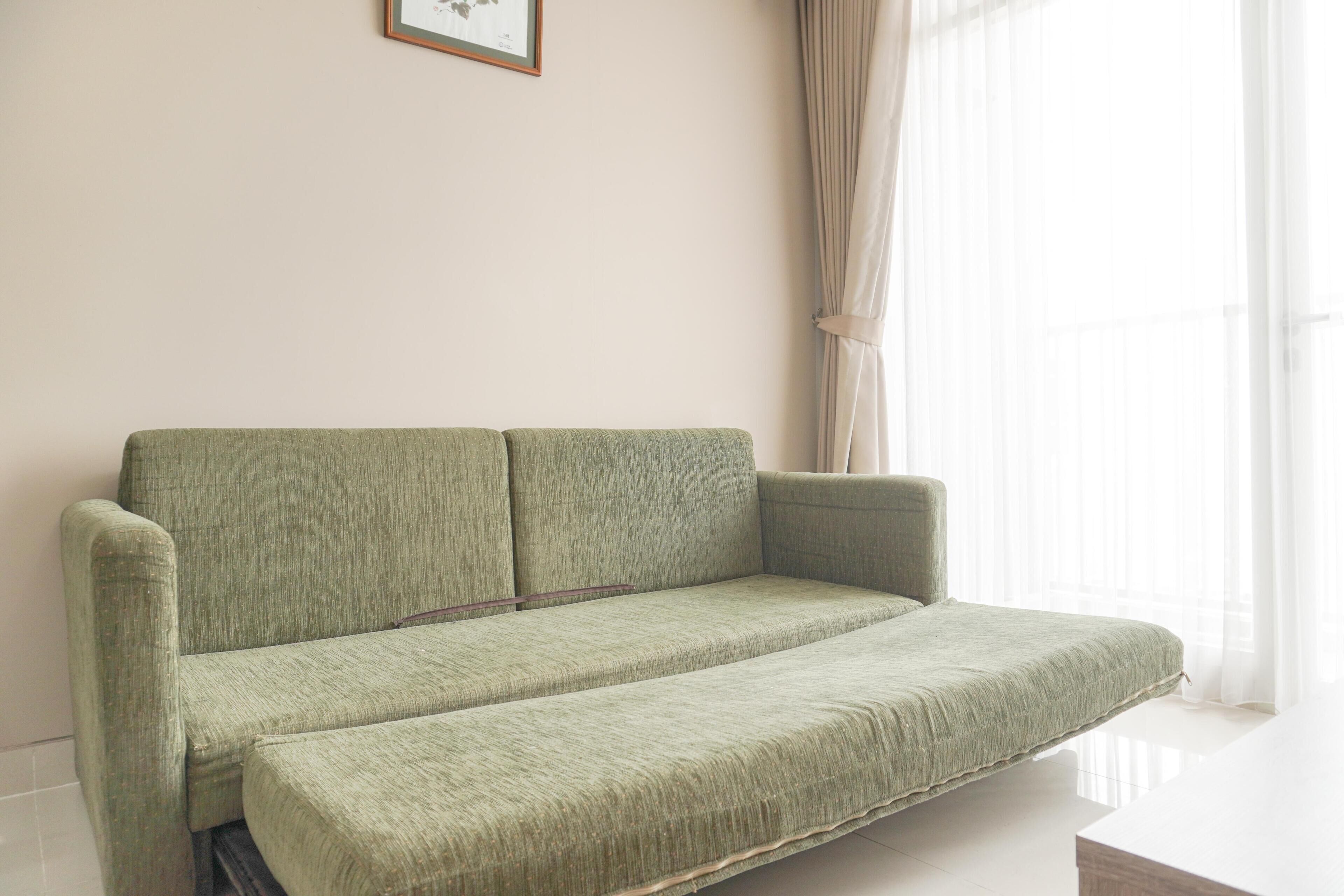 A green sofa in a room with a white wall and a window with sheer curtains.