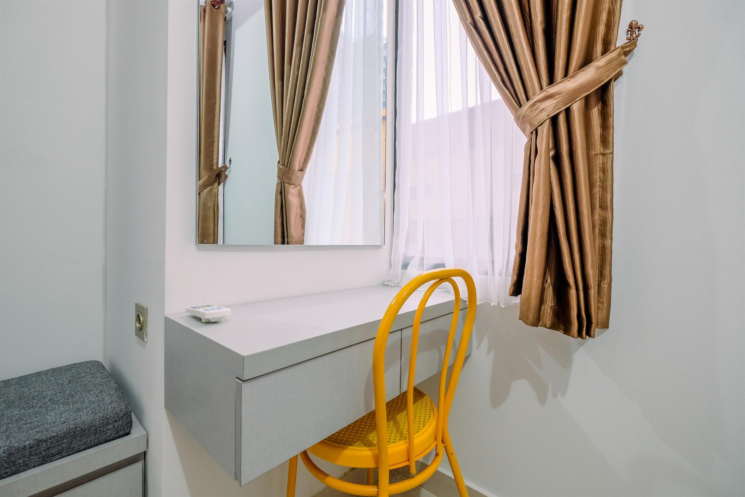 A room with a white desk, a yellow chair, a window with curtains, and a mirror.