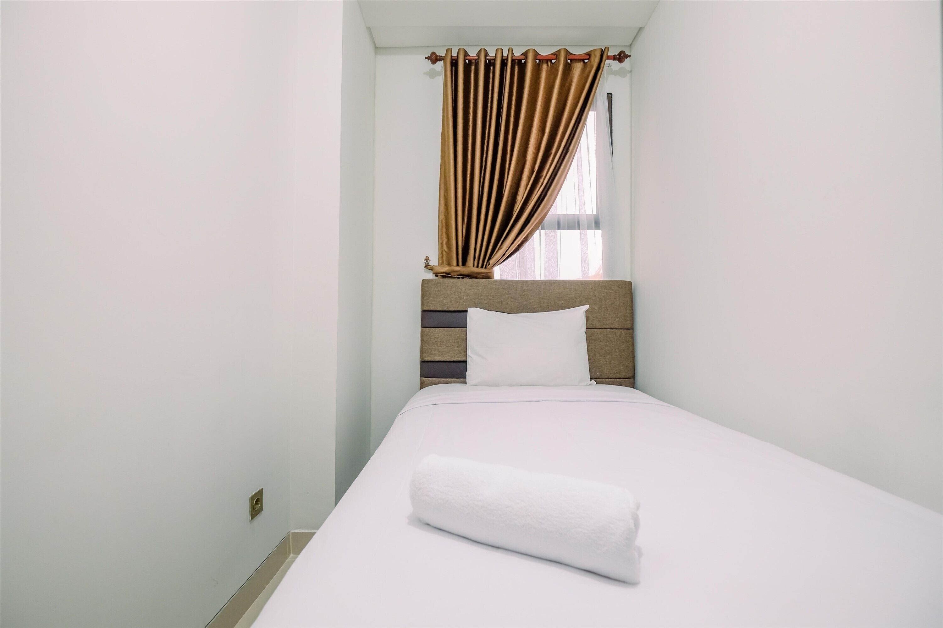 Cama bem arrumada com um edredom branco e uma toalha branca enrolada, cabeceira de madeira e uma janela com cortinas castanhas.