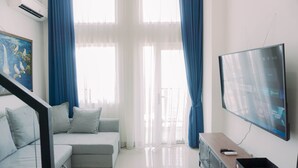 A modern living room with a grey sectional sofa, a flat-screen TV mounted on the wall, and a staircase leading to an upper level. - Comfortable And Homey Studio Loft At Kingland Avenue Apartment (South Tangerang)