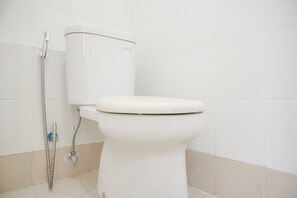 A white toilet with a tank and a shower attachment in a bathroom with white and beige tiles. - Good Choice And Minimalist 1Br Bassura City Apartment (Jakarta)