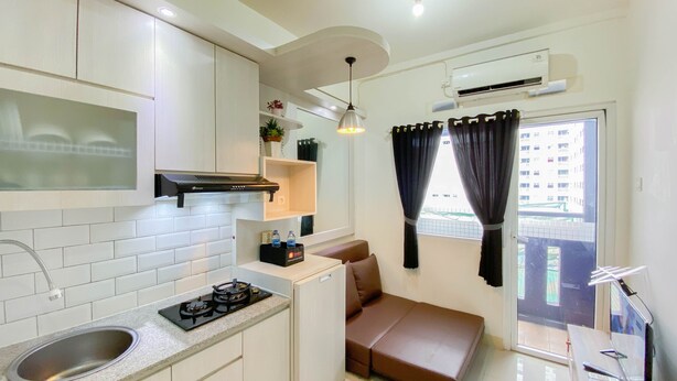 A compact kitchen with a sink, stove, and cabinets, a small seating area with a sofa, and a window with curtains. - Good Choice And Homey 2Br At Green Pramuka City Apartment (Jakarta)