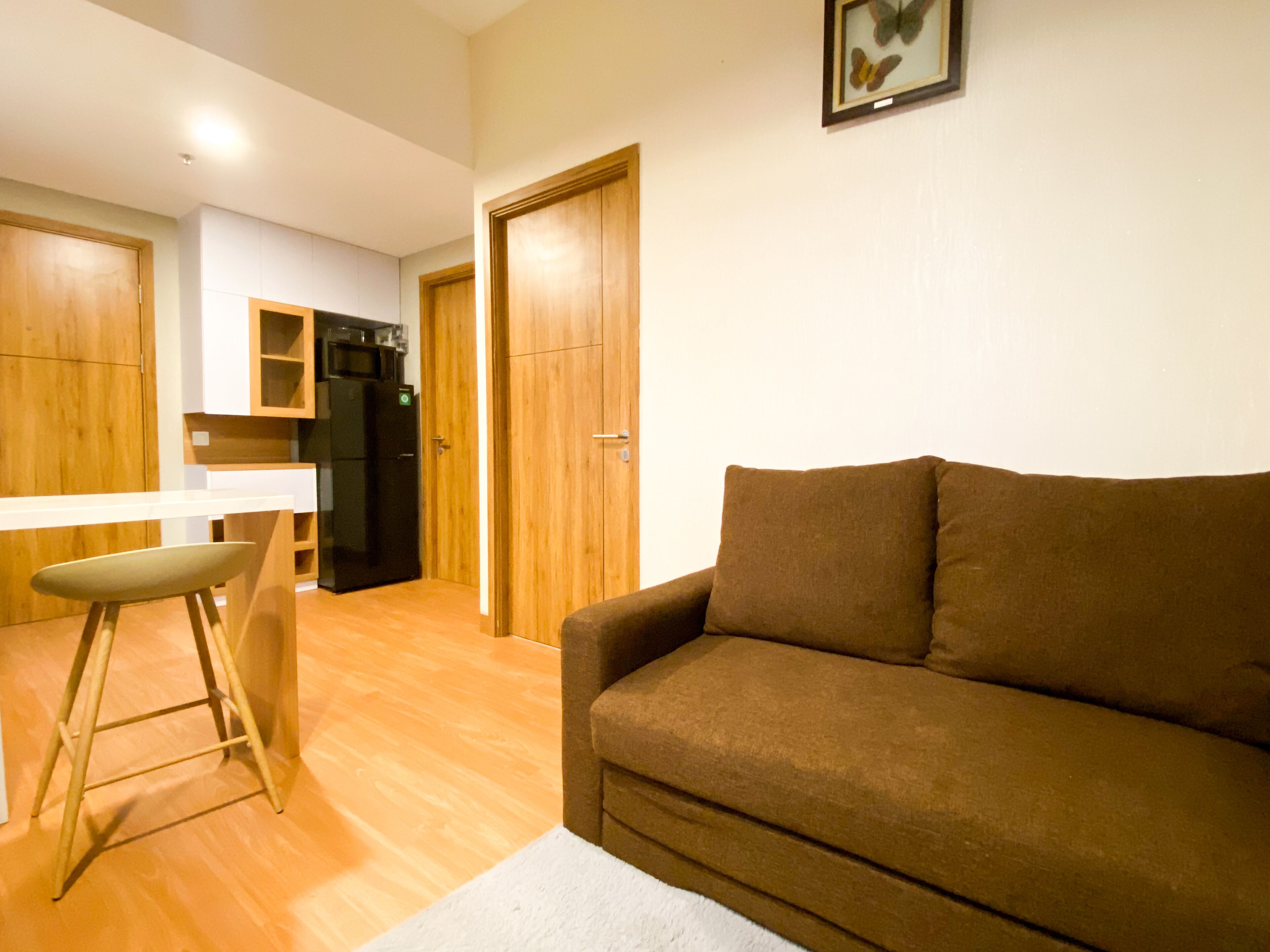 A living room with a brown sofa, a white chair, and a wooden floor. There is a kitchen area with wooden cabinets and a refrigerator.