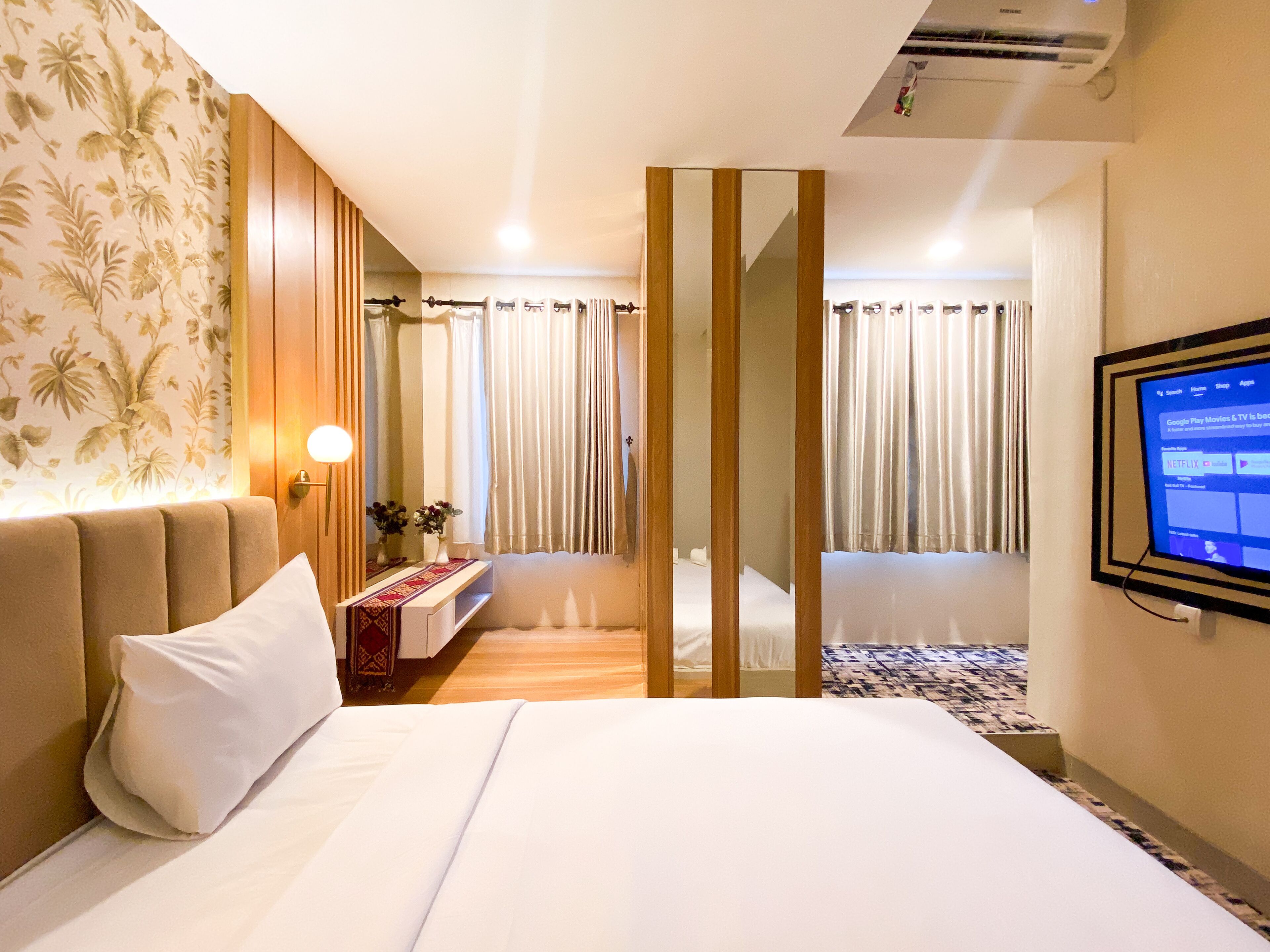 A hotel room with a bed, a bedside table with a lamp, a mirror, a TV mounted on the wall, and a window with curtains.