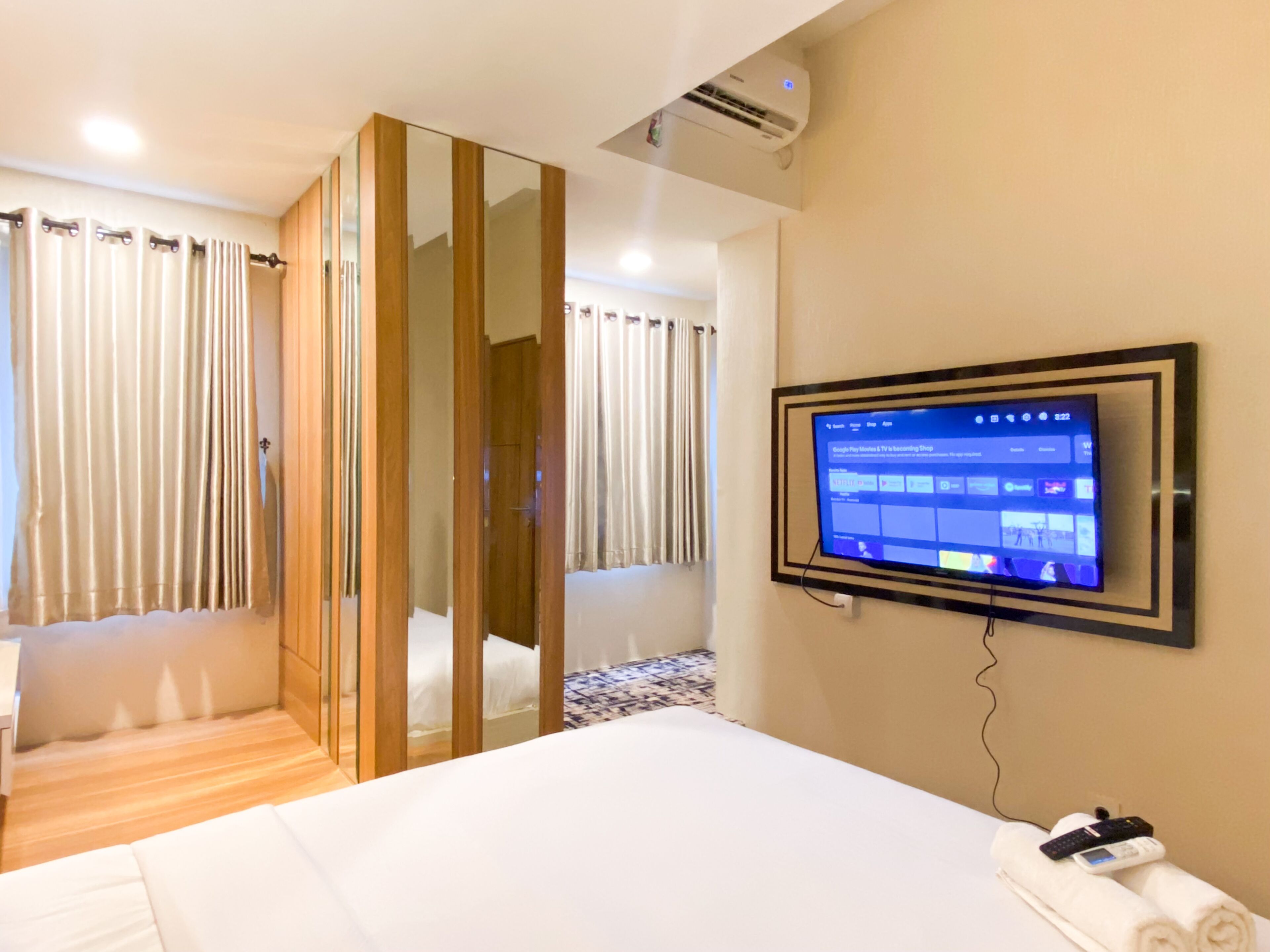 A hotel room with a bed, a TV mounted on the wall, a wooden wardrobe, and a bathroom with a shower curtain.