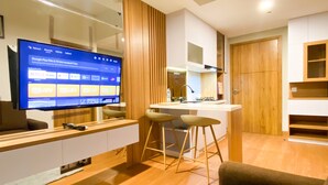 A modern living room with a flat-screen TV, wooden flooring, a kitchen area, and a wooden door. - Comfy and Modern Look 1BR LRT City Jatibening Apartment (Bekasi)