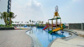 Relax by the pool surrounded by lush greenery and a children's play area with slides and climbing elements.