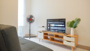 Een woonkamer met een bank, een houten tv-meubel met een flatscreen-tv waarop een Netflix-interface wordt weergegeven, een ventilator en een potplant.