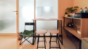 Une petite pièce moderne avec une table en bois et deux tabourets, une chaise pliante verte, une étagère avec une plante et des bouteilles d’eau, et une porte-fenêtre coulissante.