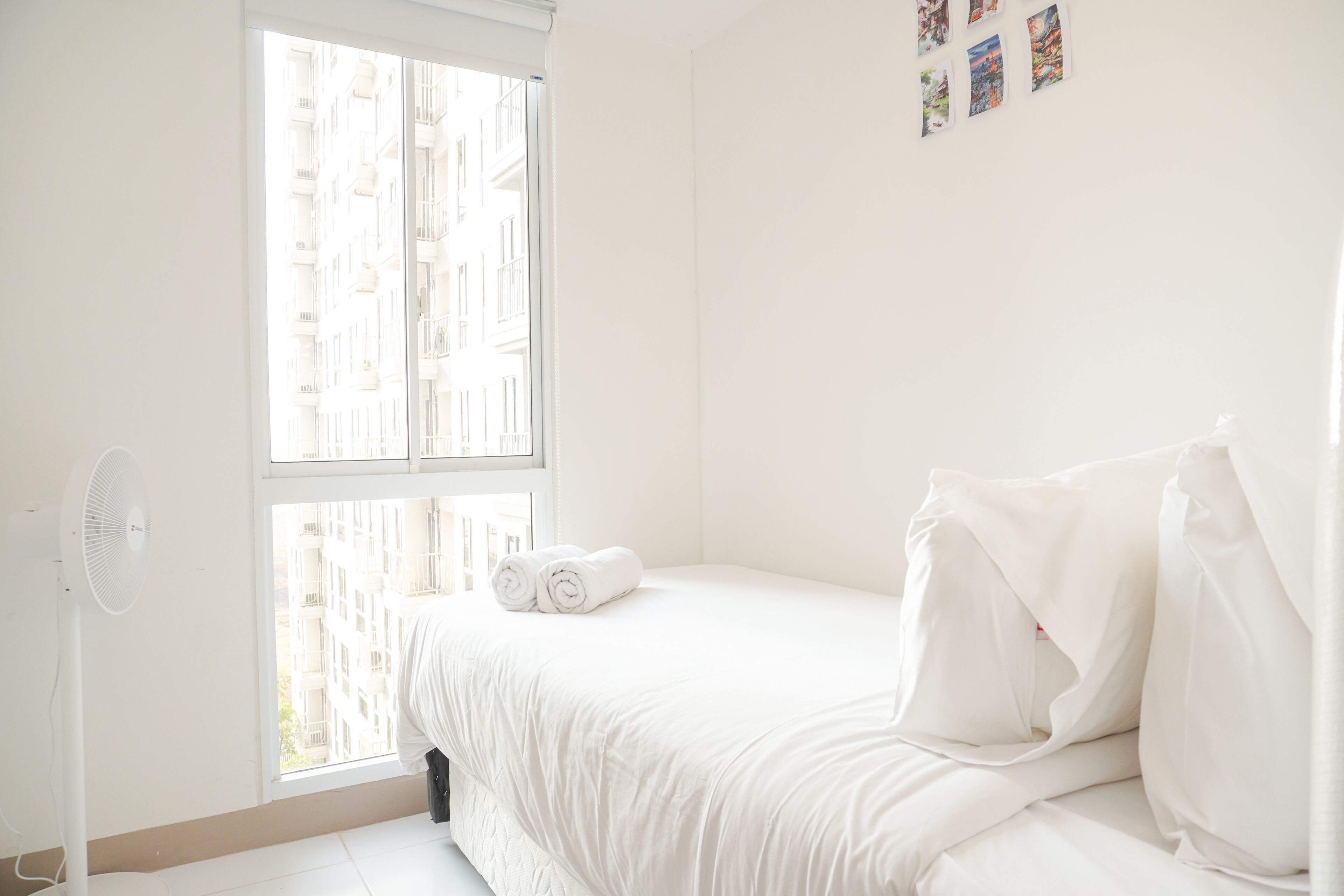 A white room with a bed, a standing fan, and a window with a view of buildings.