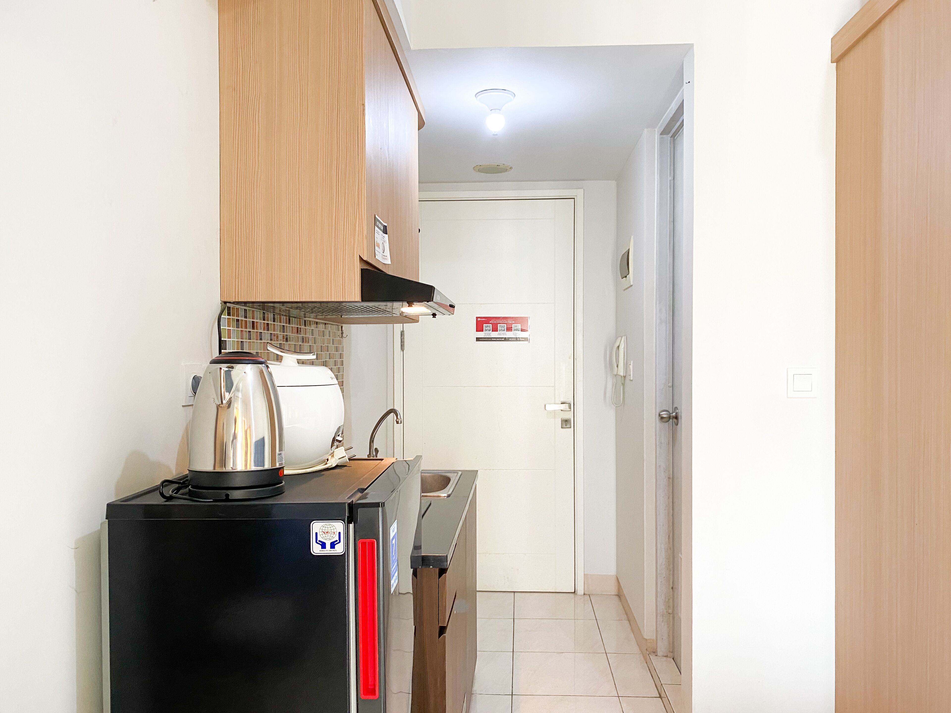 Eine kompakte Küche mit einem schwarzen Kühlschrank, einer Kaffeemaschine und einer Mikrowelle. Über der Arbeitsfläche befindet sich ein Holzschrank und im Hintergrund eine weiße Tür.