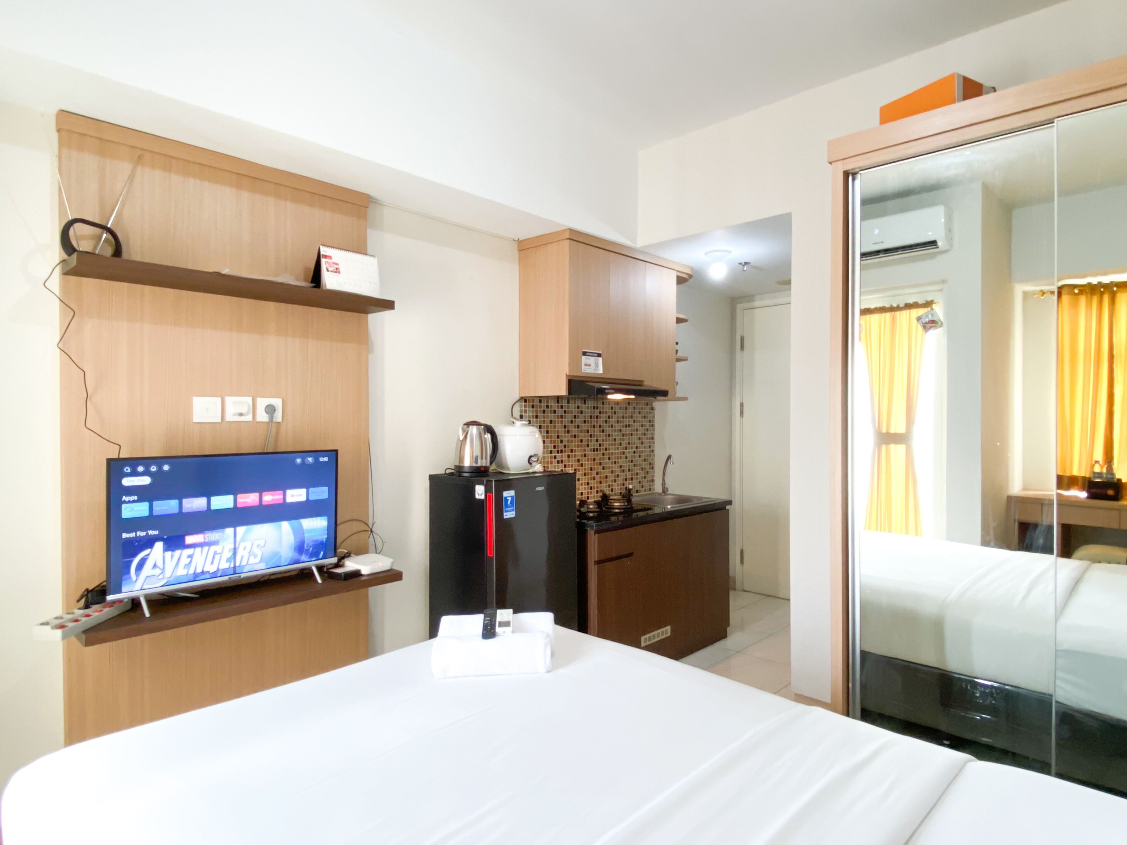 Habitació d'hotel amb un llit, una televisió que mostra 'Avengers', una zona de cuina amb nevera, fogó i microones, i un bany amb mirall i cortines.